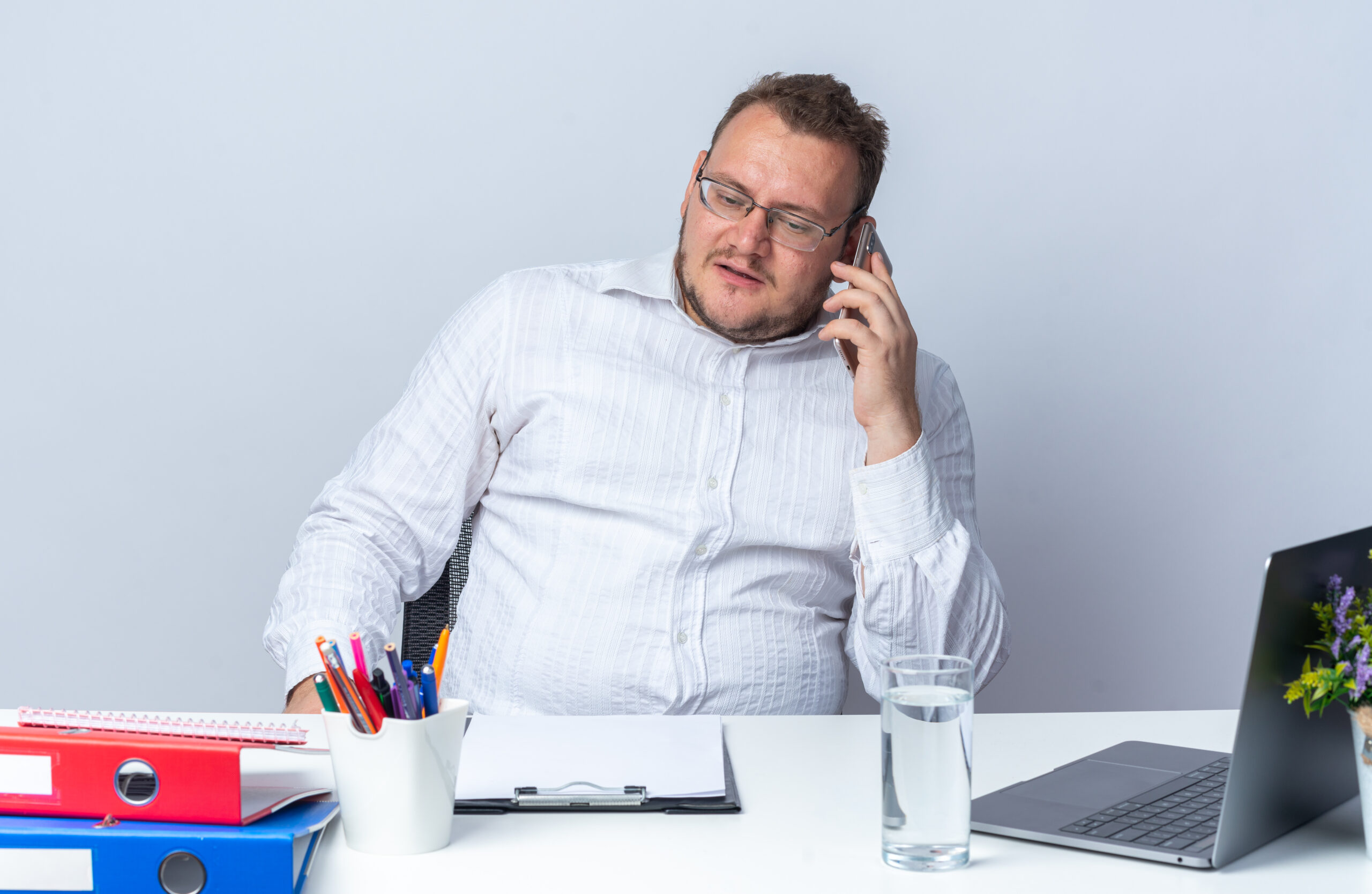 Otyły mężczyzna siedzacy w białej koszuli przy biurku i rozmawiający przez telefon.